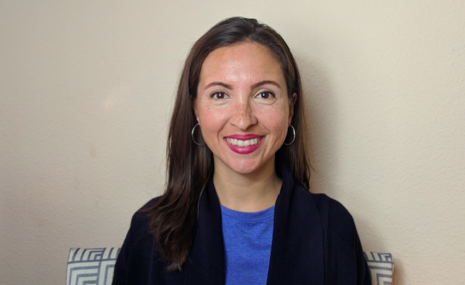 Photo of Dr. Morghan Velez wearing a blue shirt and black suit jacket, smiling, seated on a blue and white chair in front of a beige wall