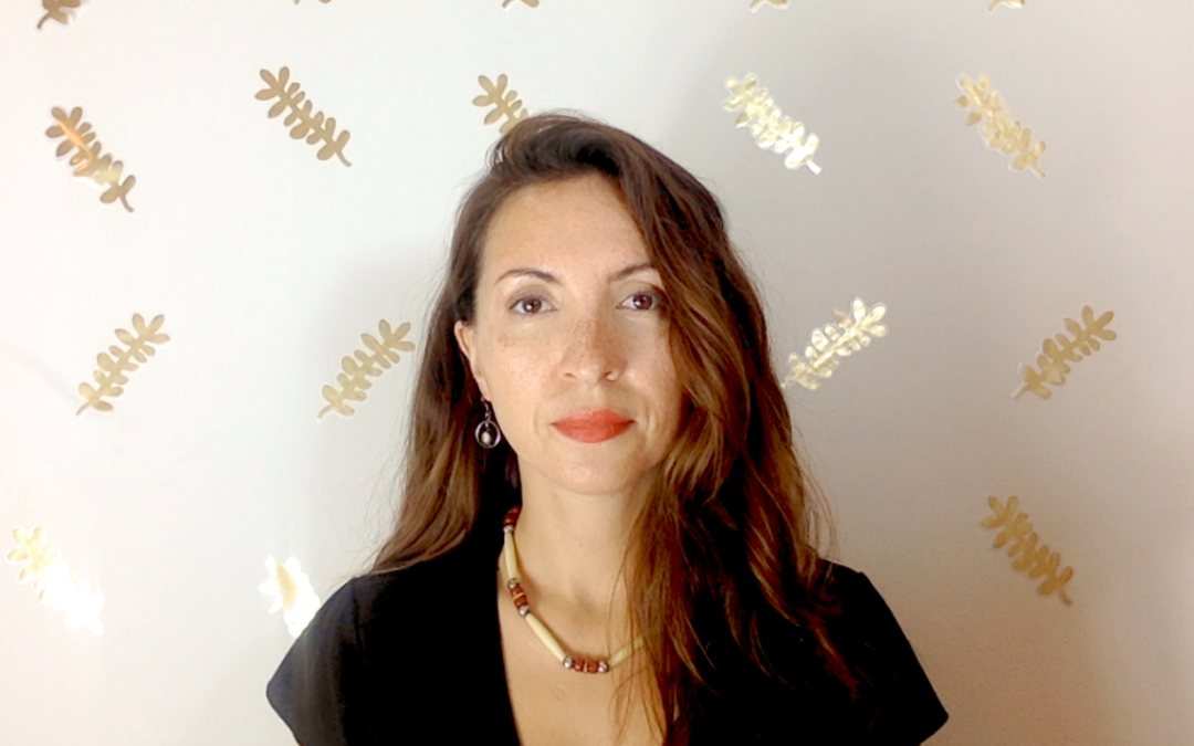 Photo of Morghan Velez Young. She is a petite woman with long, dark hair. She is wearing a black shirt and is standing in front of a white background with gold leaves on it.